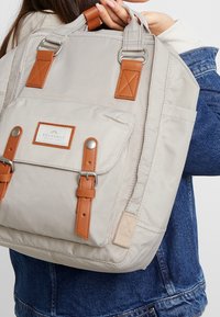 Beżowy plecak materiałowy z brązowymi skórzanymi akcentami i metalowymi elementami, z przednią kieszenią i paskami na ramię.