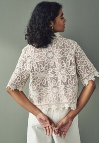 Bluse i blonde i off-white med indviklede blomster mønstre, korte ærmer og bølget kant. Model står med hænderne bag ryggen.