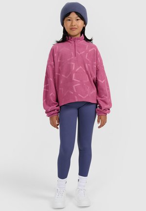 Dziecko ubrane w fioletową czapkę, różowy wzorzysty polarowy sweter, niebieskie legginsy, białe skarpetki i białe trampki, stojące na jednolitym tle.