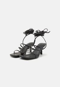 Svarta strappy högklackade sandaler med tunna läderspännen, fyrkantig tådesign och en liten klack med dekorativa snörningar vid ankeln.