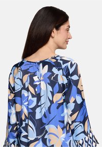 Blouse fleurie en bleu, crème et marine avec un col rond, un dos froncé et des manches longues aux ourlets volantés.