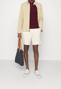 Man som bär en beige jacka över en vinröd polotröja, krämfärgade shorts, grå sneakers och håller en marinblå toteväska med bruna läderhandtag.