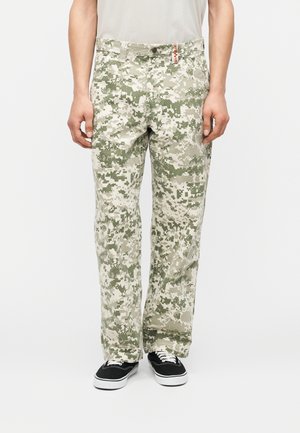 Osoba oblečená v voľne strihaných maskáčových nohaviciach a čiernych teniskách, stojaca pred bielym pozadím.