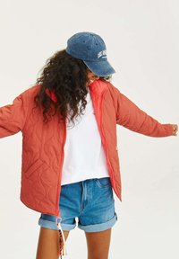 Pikowana koralowa kurtka z zamkiem błyskawicznym, bocznymi kieszeniami i kontrastową podszewką, noszona na białej koszuli i dżinsowych szortach. Niebieska czapka z napisem.