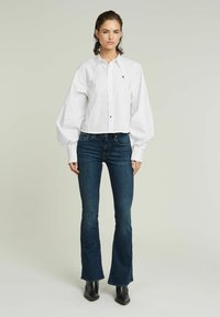 Valkoinen cropped-button-up-paita, jossa on ylisuuria hihoja, yhdistetty tummansinisisiin levennettyihin farkkuihin ja mustiin nilkkureihin. Yksinkertainen muotoilu ilman kuvioita.