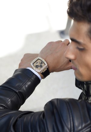 Homme en veste en cuir noire regardant une grande montre carrée en or avec un bracelet noir à son poignet.
