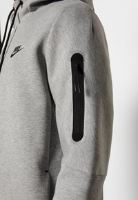 Grå sweatshirt med en svart Nike-logotyp, med en ficka med dragkedja på vänster ärm och textil med struktur. Snörning på huvan ingår.