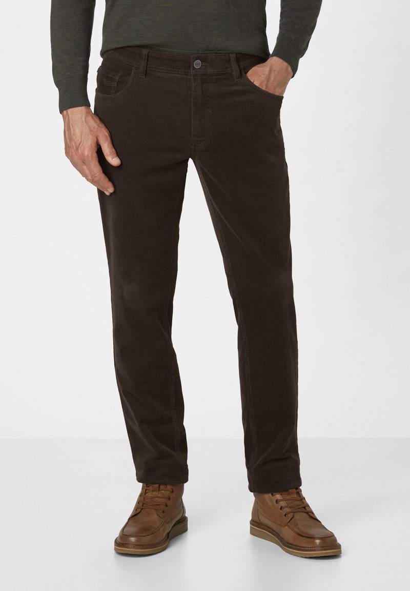 Mann in dunkelbraunen Cordhosen, mit einer Hand in der Tasche und braunen, geschnürten Lederstiefeln, steht vor einem schlichten Hintergrund.