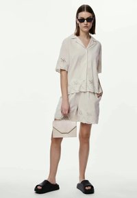 Ανοιχτό μπεζ μπλούζα κοντομάνικη με λεπτομέρειες από διαφάνεια, συνδυασμένη με κοντές σορτς της ίδιας απόχρωσης. Κρατά μια μικρή, δομημένη τσάντα και φορά μαύρα πέδιλα.