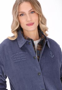 Blondynka o niebieskich oczach ma na sobie niebieską kurtkę z sztruksu na brązowej i niebieskiej koszuli w kratę, delikatnie się uśmiechając na białym tle.