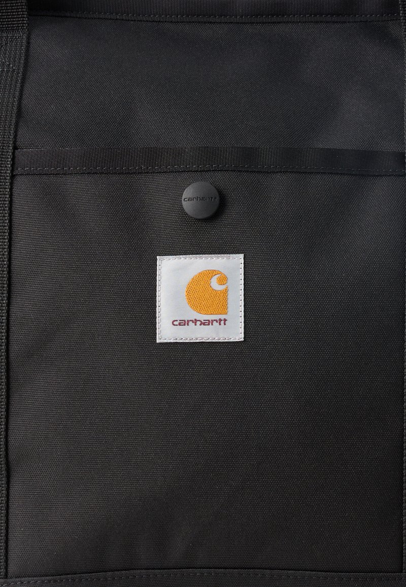 Zwarte stoffen tas met een voorvak dat is bevestigd met een zwarte knoop, met een witte patch met een oranje Carhartt-logo en rode tekst.