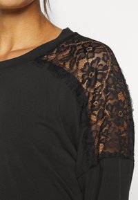 Maglietta nera a maniche lunghe con dettagli in pizzo sulle spalle. Il pizzo presenta un motivo floreale con bordi frangiati, in contrasto con il tessuto solido.