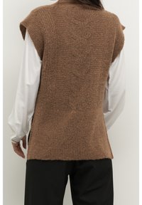 Persoon die een gebreide bruine mouwloze trui draagt over een wit shirt met lange mouwen en een zwarte broek, gezien van achteren tegen een effen achtergrond.