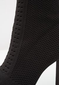 Gros plan sur une bottine à talon haut noire en maille texturée avec de petites perforations et un détail de rayure verticale.
