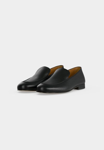 Paire de chaussures en cuir noir à enfiler avec petits talons et bouts arrondis sur un fond clair uni.