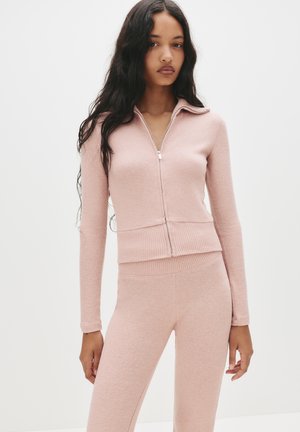 Roza zip-up dukserica s rebrastim pojasom i rukavima, u kombinaciji s uskim hlačama u istom tonu. Mekana tekstura, lagani materijal. Minimalistički dizajn.