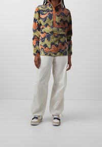 Fleecová mikina s vícebarevnými vlnkovými vzory ve zelené, oranžové a modré barvě, s hnědým límcem na knoflíky a přední kapsou, doplněná o bílé kalhoty.