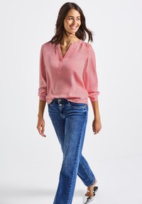 Blusa a maniche lunghe rosa chiaro con scollatura a V, abbinata a jeans blu. La blusa ha una vestibilità comoda e una texture morbida. I dettagli includono le maniche arrotolate.