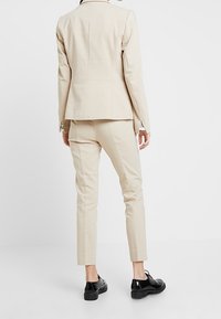 Femme portant un blazer beige ajusté et un pantalon slim assorti, associé à des chaussures derby en cuir verni noir, se tenant devant un fond blanc.