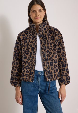 Femme portant une veste zippée imprimé léopard marron et noir, un haut blanc et un jean bleu, debout devant un fond uni.