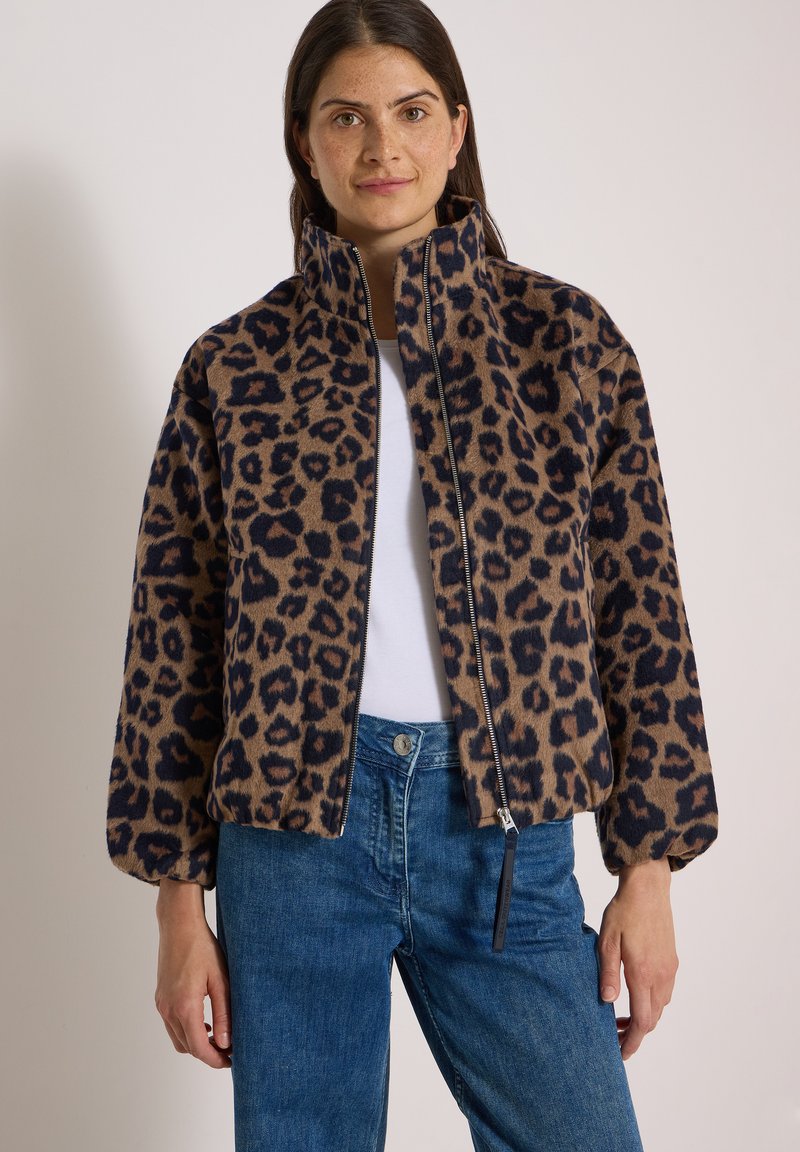 Femme portant une veste zippée imprimé léopard marron et noir, un haut blanc et un jean bleu, debout devant un fond uni.