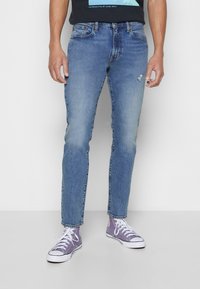 Světlé modré džíny z denimu s rovný střihem, mírným opotřebením na koleni a klasickým designem s pěti kapsami. Nošeny s fialovými teniskami.