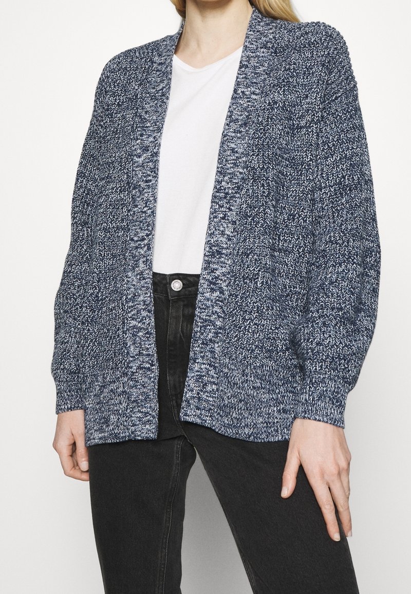 Personne portant un cardigan bleu et blanc en grosse maille à ouverture sur le devant, par-dessus une chemise blanche et un jean noir, debout devant un fond uni.