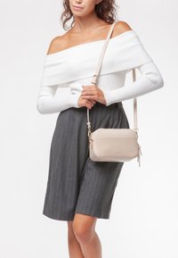 Bolso cruzado beige hecho de material suave, con forma rectangular, herrajes dorados y cierre de cremallera con detalle de borlas.
