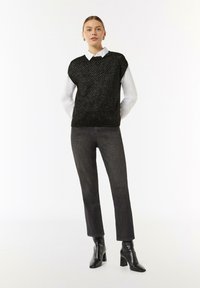 Czarny bezrękawnik z fakturowanym wzorem, noszony na białej koszuli z kołnierzykiem, w zestawieniu z ciemnymi dżinsami i czarnymi botkami na obcasie.