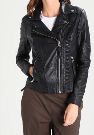 Veste de motard en cuir noir avec fermeture éclair diagonale, poches zippées et détails matelassés portée sur une chemise blanche et un pantalon marron.