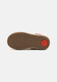 Semelle de chaussure en caoutchouc brun avec un motif de grip texturé, un logo circulaire et un emblème de basketball rouge. Design plat, bords lisses, matériau durable.