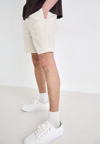 Beżowe lniane szorty z bocznymi kieszeniami, o luźnym kroju. Noszone z białymi sneakersami i jasnoszarymi skarpetkami. Gładka tekstura, minimalistyczny design.