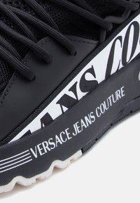 Μαύρο αθλητικό παπούτσι με επάνω μέρος από δίχτυ, λαστιχένιες λεπτομέρειες, εξέχον λευκό λογότυπο και υφασμένο πέλμα με κυματιστό σχέδιο.