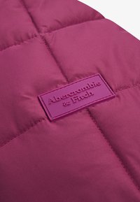 Jachetă pufoasă într-o nuanță profundă de fucsia, cu secțiuni matlasate. Prezintă un patch din cauciuc texturat, de culoare purpurie, cu inscripția "Abercrombie & Fitch."