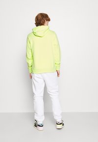 Felpa con cappuccio verde neon brillante, vestibilità comoda, tasca a marsupio e cappuccio. Abbinata a pantaloni della tuta bianchi e sneakers multicolore.