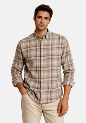 Jeune homme portant une chemise à carreaux beige et marron boutonnée avec les manches retroussées et un pantalon beige clair, debout avec une main dans la poche.