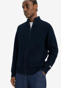 Cárdigan azul marino de canalé con cremallera y cuello alto, dos bolsillos frontales y puños ajustados. Fabricado en un tejido de punto grueso y texturizado.
