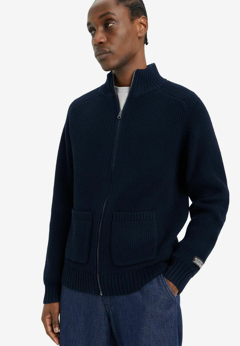Cárdigan azul marino de canalé con cremallera y cuello alto, dos bolsillos frontales y puños ajustados. Fabricado en un tejido de punto grueso y texturizado.