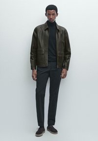 Bruine leren jas met een staande kraag, twee voorzakken, gecombineerd met een zwarte coltrui en donkere broek, tegen een neutrale achtergrond.