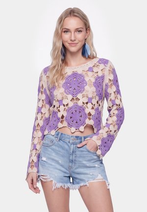Giovane donna con capelli biondi lunghi che indossa un crop top lavorato a uncinetto viola e beige con maniche lunghe e pantaloncini di jeans strappati azzurri.