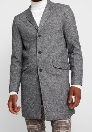 Homme portant un manteau gris texturé boutonné sur un col roulé blanc et un pantalon à carreaux, debout devant un fond uni.