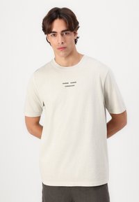 Camiseta beige clara hecha de algodón suave, con mangas cortas y cuello redondo. En el frente tiene el texto negro "SAMSØE SAMSØE COPENHAGUE."