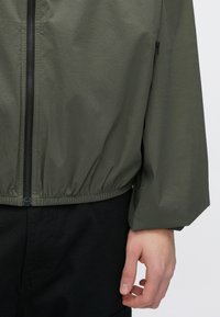 Ελαφρύ μπουφάν σε ελιά πράσινο χρώμα με ελαστικό τελείωμα και μανσέτες, διαθέτει κεντρικό φερμουάρ και πλαϊνή τσέπη με διακριτική λεπτομέρεια φερμουάρ.