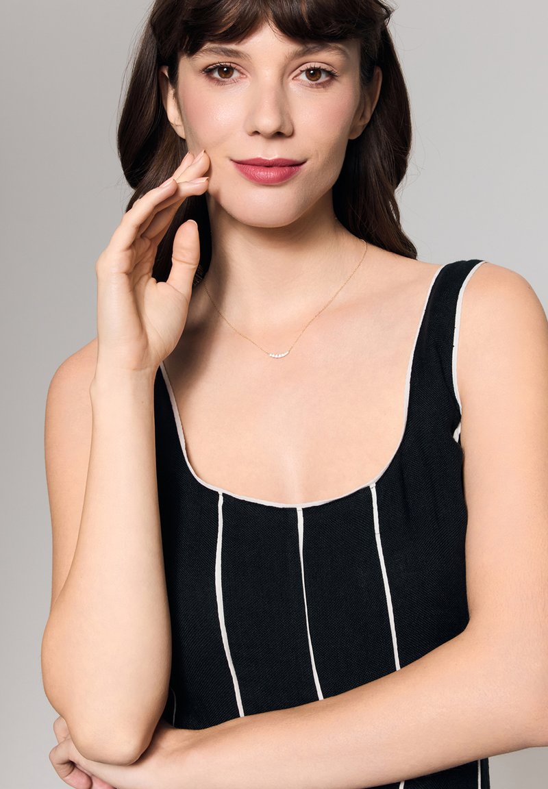 Czarny top bez rękawów z białymi paskami i szerokim dekoltem. Modelka nosi delikatny złoty naszyjnik z małymi akcentami i pozując trzyma rękę na policzku.