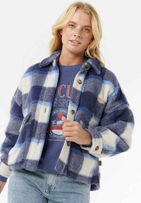 Blonde vrouw die een blauw-wit geruite fleece jack draagt over een grafisch blauw sweatshirt en lichtblauwe spijkerbroek.