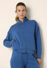 Felpa blu con zip in un tessuto morbido, dotata di un collo alto, maniche lunghe e sottili dettagli decorativi lungo la zip.