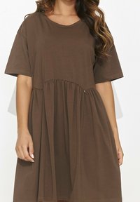 Femme portant une robe courte à manches courtes, ample et marron, avec un détail froncé à la taille, se tenant devant un fond blanc uni.