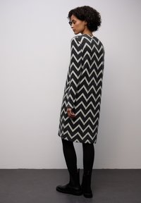 Pletené šaty se vzorem cikcak v černé a bílé barvě, s dlouhými rukávy, do poloviny lýtek, nošené přes černé punčocháče a kotníkové boty.