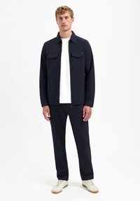 Marineblauwe jas met twee borstzakken, gecombineerd met marineblauwe broek. Materiaal lijkt glad; gestyled met een wit T-shirt en lichte sneakers.