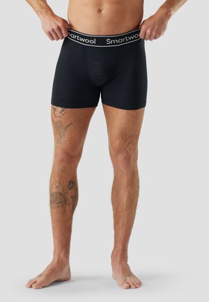 Vita a piedi di uomo che indossa boxer Smartwool neri, mano destra che regola la vita, tatuaggi visibili sulla gamba sinistra e sui piedi su sfondo uniforme.